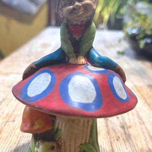 May include: A ceramic gnome figurine sitting on a red and blue mushroom with white polka dots. The mushroom has a green base with a yellow caterpillar and a smaller red mushroom.