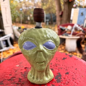 May include: A green alien head ceramic pipe with a wooden top. The alien has large blue eyes and a small mouth.