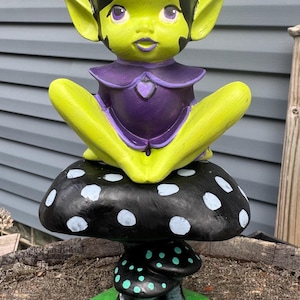 May include: A whimsical ceramic figurine of a seated fairy with a green complexion, black hair, and purple accents. The fairy is perched atop a black mushroom with white polka dots, with additional smaller mushrooms at the base. The base is green.