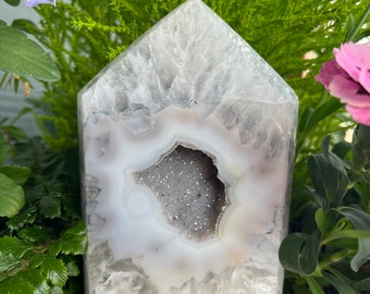 Pode incluir: Um cristal de ágata branco com uma grande geoda aberta. A geoda está cheia de cristais menores e brilhantes. O cristal tem a forma de uma casa e está sentado sobre uma base de madeira.