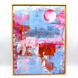 May include: Abstract painting with a pink circle in the upper right corner. The painting is made up of layers of blue, pink, and orange paint. The painting is framed in a light wood frame.