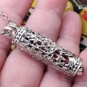 May include: Silver-colored pendant necklace with a cylindrical vial design. The vial features intricate filigree and small clear gemstones. The necklace is suspended from a delicate chain. The brand name "BloodVials" is visible in the upper right corner.