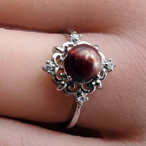 May include: A silver ring featuring a dark red, round gemstone at the center, surrounded by a decorative filigree design and small, clear accent stones. The ring is on a finger.