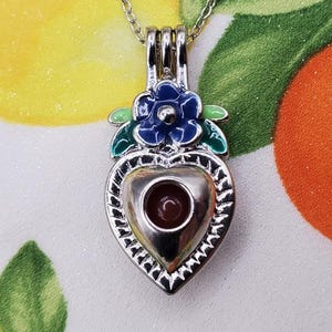K&ouml;nnte beinhalten: Eine silberfarbene herzf&ouml;rmige Anh&auml;ngerkette mit einem kleinen, runden, roten Stein im Inneren. Der Anh&auml;nger ist mit einer blauen Blume und gr&uuml;nen Bl&auml;ttern verziert. Die Kette tr&auml;gt den Text "Blood Vials" oben.
