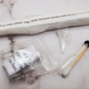 K&ouml;nnte beinhalten: Eine Auswahl an Artikeln, darunter eine Packung mit Text, eine Spritze, Wattest&auml;bchen und andere kleine Komponenten. Der Text auf der Packung lautet: "Squirt glue into silver cap, and remove excess with Q-tip to limit coverage on".