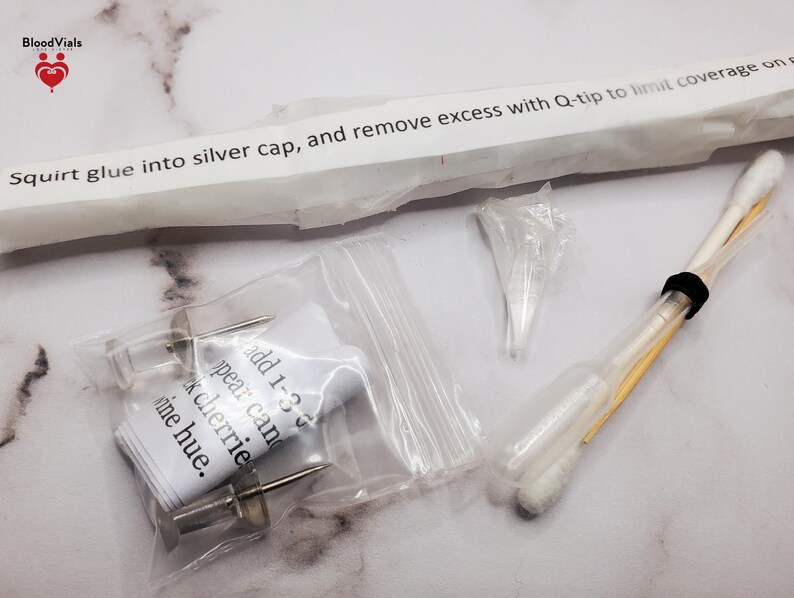 K&ouml;nnte beinhalten: Eine Auswahl an Artikeln, darunter eine Packung mit Text, eine Spritze, Wattest&auml;bchen und andere kleine Komponenten. Der Text auf der Packung lautet: "Squirt glue into silver cap, and remove excess with Q-tip to limit coverage on".