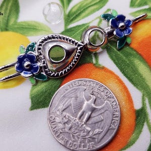 K&ouml;nnte beinhalten: Ein silberfarbener Flaschenverschluss mit herzf&ouml;rmigem Design, verziert mit blauen und gr&uuml;nen floralen Akzenten. Der Verschluss wird neben einer US-Viertel-Dollar-M&uuml;nze gezeigt. Das BloodVials-Logo befindet sich oben links.