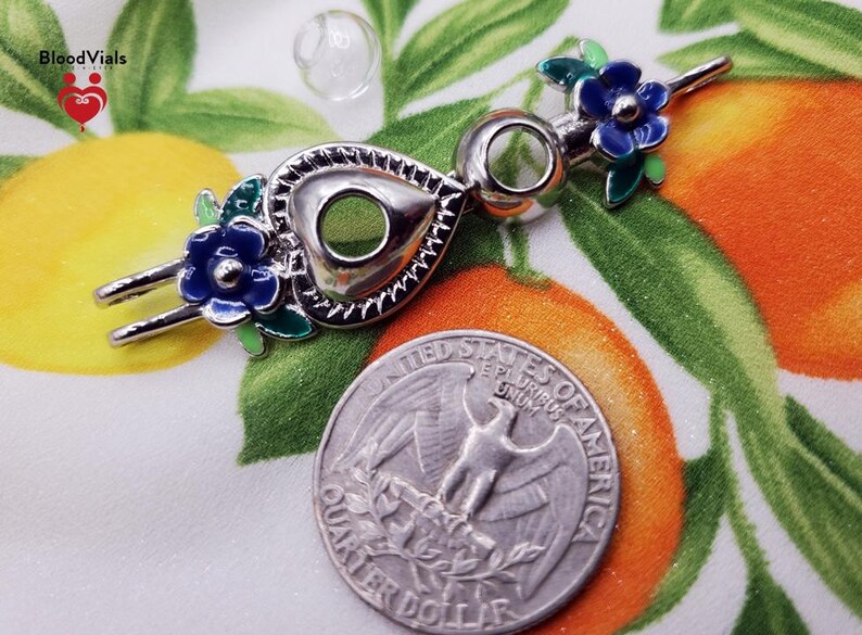 K&ouml;nnte beinhalten: Ein silberfarbener Flaschenverschluss mit herzf&ouml;rmigem Design, verziert mit blauen und gr&uuml;nen floralen Akzenten. Der Verschluss wird neben einer US-Viertel-Dollar-M&uuml;nze gezeigt. Das BloodVials-Logo befindet sich oben links.