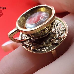 May include: A gold-colored ring featuring a miniature teacup and saucer design. The teacup holds a clear, oval-shaped stone with red inclusions. The saucer includes a tiny spoon. The ring is held by a finger against a red background.