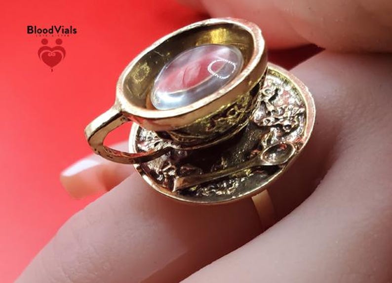 May include: A gold-colored ring featuring a miniature teacup and saucer design. The teacup holds a clear, oval-shaped stone with red inclusions. The saucer includes a tiny spoon. The ring is held by a finger against a red background.