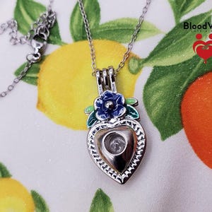 K&ouml;nnte beinhalten: Eine silberfarbene herzf&ouml;rmige Anh&auml;nger-Halskette mit einer blauen Blume und gr&uuml;nen Bl&auml;ttern. Der Anh&auml;nger hat eine klare, hohle Mitte. Die Halskette befindet sich auf einem Hintergrund mit Zitronen und Orangen. Der Text "Blood Vials" ist sichtbar.