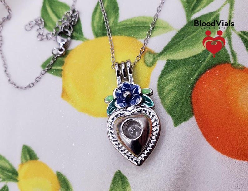 K&ouml;nnte beinhalten: Eine silberfarbene herzf&ouml;rmige Anh&auml;nger-Halskette mit einer blauen Blume und gr&uuml;nen Bl&auml;ttern. Der Anh&auml;nger hat eine klare, hohle Mitte. Die Halskette befindet sich auf einem Hintergrund mit Zitronen und Orangen. Der Text "Blood Vials" ist sichtbar.