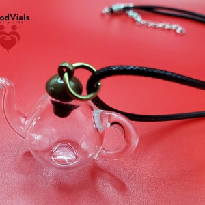 May include: A clear glass teapot pendant necklace with a black cord. The teapot has a small handle and spout, and a dark brown cap. The necklace is on a red surface. The brand name "BloodVials" is visible in the top left corner.