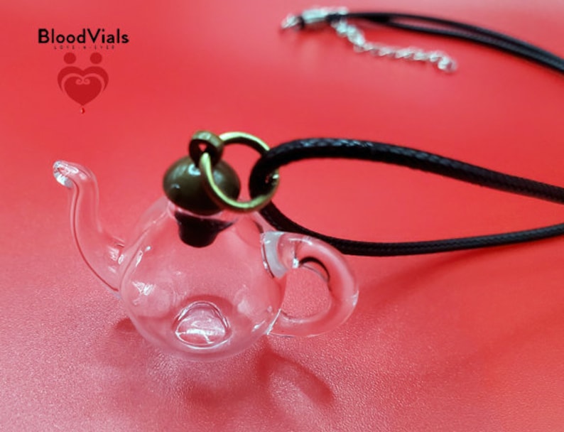 May include: A clear glass teapot pendant necklace with a black cord. The teapot has a small handle and spout, and a dark brown cap. The necklace is on a red surface. The brand name "BloodVials" is visible in the top left corner.
