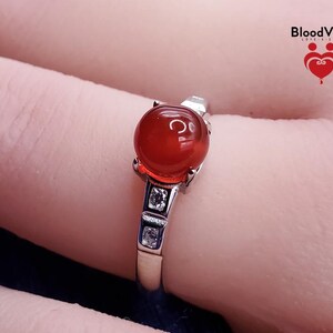May include: A silver ring featuring a large, round, reddish-orange gemstone. The ring has a decorative setting with small, square, clear stones. The brand name "BloodVials" is visible in the upper right corner.