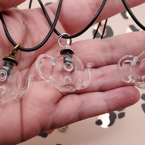 May include: Three clear glass teapot pendants on black cords. Each pendant has a different colored cap: bronze, silver, and gold. The pendants are displayed against a blurred background with the "BloodVials" logo.