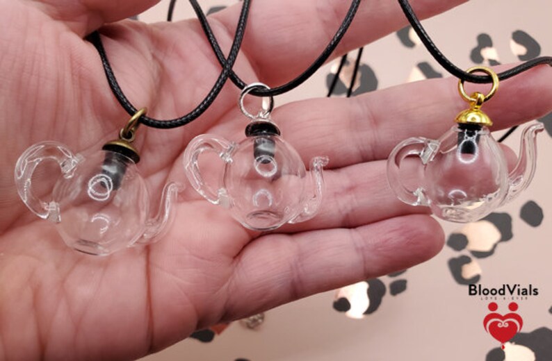 May include: Three clear glass teapot pendants on black cords. Each pendant has a different colored cap: bronze, silver, and gold. The pendants are displayed against a blurred background with the "BloodVials" logo.