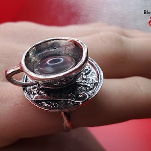May include: A silver-colored ring featuring a miniature teacup and saucer design. The teacup is filled with a clear liquid. The ring is worn on a finger. The brand name "BloodVials" is visible in the upper right corner.