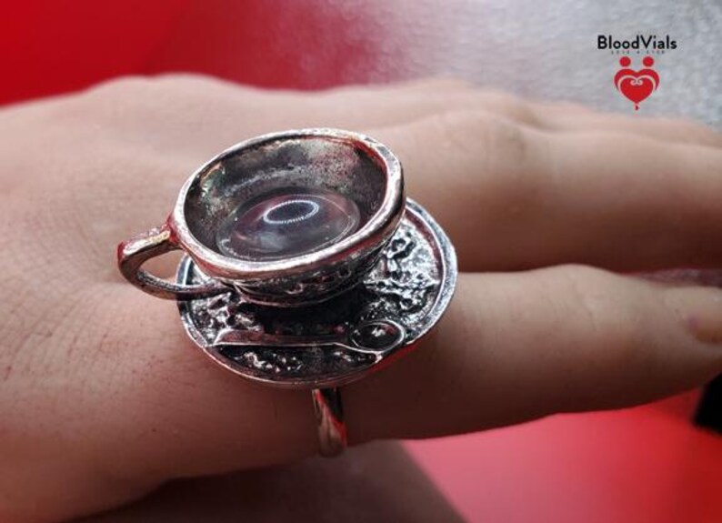 May include: A silver-colored ring featuring a miniature teacup and saucer design. The teacup is filled with a clear liquid. The ring is worn on a finger. The brand name "BloodVials" is visible in the upper right corner.