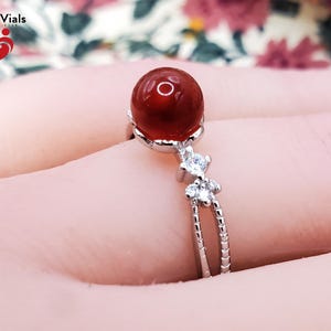 May include: A silver ring featuring a large, round, red gemstone. The ring has a split band with small, clear accent stones. The ring is set against a soft pink background. The brand name "Blood Vials" is visible in the upper left corner.