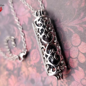 May include: A silver-colored pendant necklace with a cylindrical, filigree-style pendant. The pendant features heart-shaped cutouts and a dark interior. The necklace chain is also silver. The words "BloodVials" are in the top left corner.