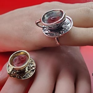 May include: Two rings, each featuring a miniature teacup and saucer design. One ring is silver-toned, the other gold-toned. The teacups appear to be filled with a red liquid. The rings are displayed on a hand against a red background.
