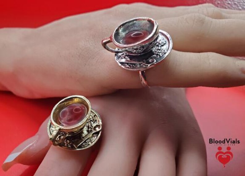 May include: Two rings, each featuring a miniature teacup and saucer design. One ring is silver-toned, the other gold-toned. The teacups appear to be filled with a red liquid. The rings are displayed on a hand against a red background.