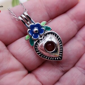 K&ouml;nnte beinhalten: Eine silberfarbene herzf&ouml;rmige Anh&auml;ngerkette mit einer blauen Blume und gr&uuml;nen Bl&auml;ttern. Der Anh&auml;nger hat einen kleinen, runden, rotbraunen Stein in der Mitte. Die Kette wird in einer Hand gehalten.