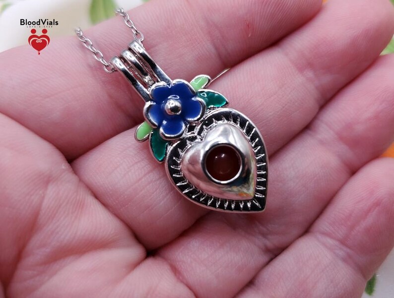 K&ouml;nnte beinhalten: Eine silberfarbene herzf&ouml;rmige Anh&auml;ngerkette mit einer blauen Blume und gr&uuml;nen Bl&auml;ttern. Der Anh&auml;nger hat einen kleinen, runden, rotbraunen Stein in der Mitte. Die Kette wird in einer Hand gehalten.