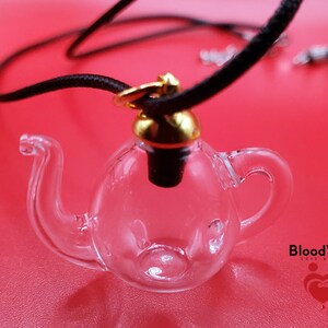 May include: A clear glass teapot-shaped vial pendant with a black cord necklace. The vial has a gold-colored cap and a black stopper. The background is a solid red color. The text "BloodVials" is visible in the lower right corner.