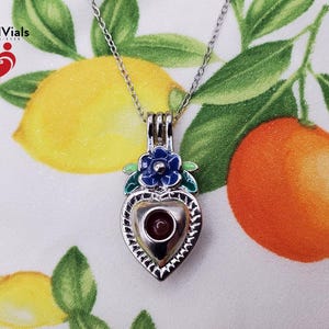 K&ouml;nnte beinhalten: Silberfarbene herzf&ouml;rmige Anh&auml;nger-Halskette mit einem kleinen, runden, roten Stein in der Mitte. Der Anh&auml;nger ist mit einer blauen Blume und gr&uuml;nen Bl&auml;ttern verziert. Der Hintergrund zeigt Zitronen und Orangen.