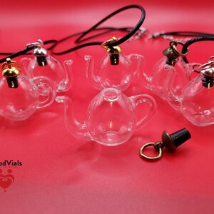 May include: Clear glass teapot-shaped pendants with black cords and metal accents. The pendants have gold, silver, and bronze-colored tops. The image is on a red background. The brand name "BloodVials" is visible in the lower left corner.