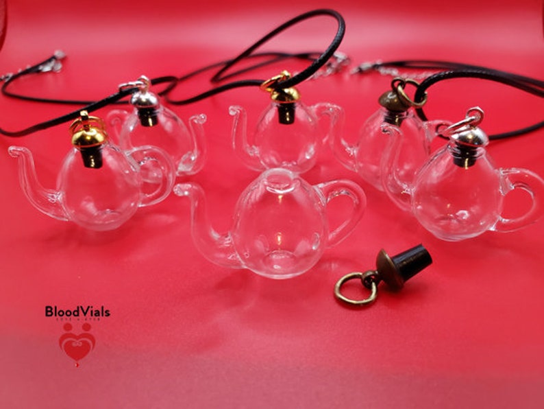 May include: Clear glass teapot-shaped pendants with black cords and metal accents. The pendants have gold, silver, and bronze-colored tops. The image is on a red background. The brand name "BloodVials" is visible in the lower left corner.