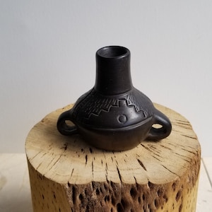 May include: A small, black ceramic vase with two handles and a geometric design. The vase is sitting on a wooden stump.