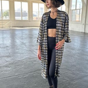 May include: A woman wearing a black and gold striped long-sleeved kimono with a black sports bra and black leggings. She is wearing a black hat and black boots.