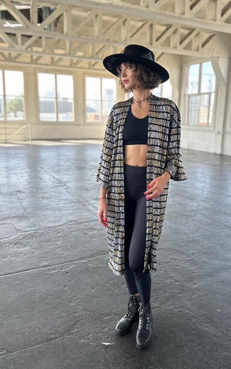 May include: A woman wearing a black and gold striped long-sleeved kimono with a black sports bra and black leggings. She is wearing a black hat and black boots.