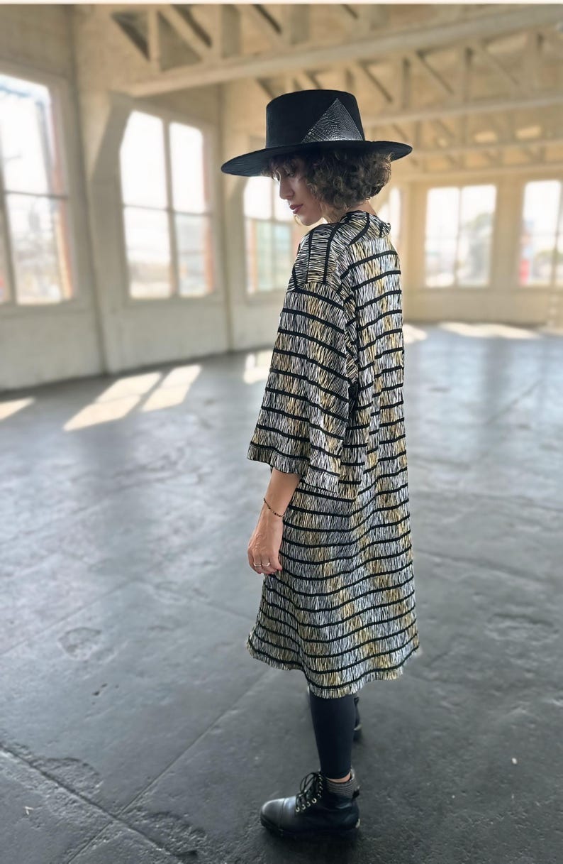 May include: A person wearing a black and white striped dress with a black hat. The dress has a loose fit and falls to the knees. The person is wearing black tights and black boots.