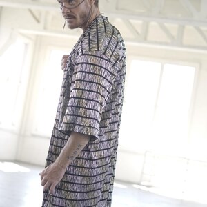 May include: A man wearing a black and gold striped long-sleeved shirt with a fringe detail. He is wearing black pants and has a tattoo on his arm.