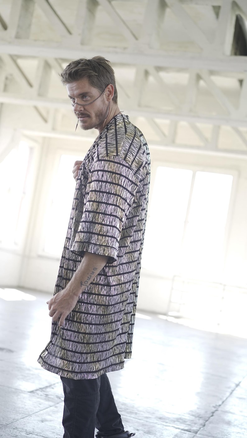 May include: A man wearing a black and gold striped long-sleeved shirt with a fringe detail. He is wearing black pants and has a tattoo on his arm.