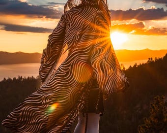 May include: A person wearing a long, black and white patterned hooded robe stands on a rocky cliff overlooking a lake and mountains at sunset. The sun is setting behind the person, creating a warm glow.