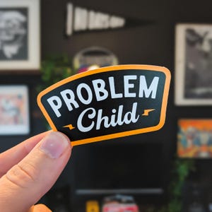 May include: A die-cut sticker with a black background and a bright orange border. The text reads "PROBLEM" in a bold, white, sans-serif font, with the word "Child" written below in a white, cursive script, flanked by two small orange lightning bolts.