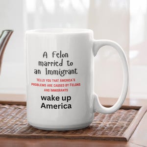 May include: White ceramic mug with the text "A felon married to an Immigrant" and "wake up America". The mug also includes the text "TELLS YOU THAT AMERICA'S PROBLEMS ARE CAUSED BY FELONS AND IMMIGRANTS". A silver teapot sits to the left.