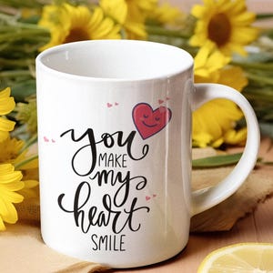 May include: A white ceramic mug featuring black cursive text that reads "you MAKE my heart SMILE" with a small red heart graphic containing a smiling face. The mug has a standard handle and a smooth finish, displaying clear, legible lettering.