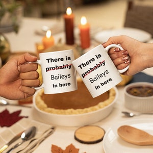 Op de afbeelding: Twee witte keramische mokken met de tekst "There is probably Baileys in here" worden omhoog gehouden voor een toast. De mokken staan op een tafel gedekt voor een maaltijd, met een taart en kaarsen op de achtergrond.