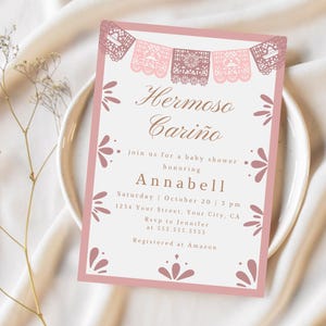 May include: Pink baby shower invitation with a festive Mexican theme. The invitation features a banner of paper cutouts with the text "Hermoso Cariño" and the words "join us for a baby shower honoring Annabell".
