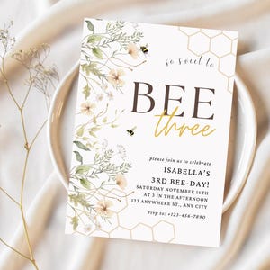 May include: A white invitation card with a bee and honeycomb design. The text reads "so sweet to BEE three" and includes details for a birthday party.