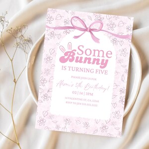 以下が含まれることがあります： 「Some Bunny Is Turning Five」の文字が入ったピンク色の誕生日招待状。招待状には花柄とピンクのリボンが付いています。招待状は、白いお皿の上に、乾燥した花の小枝と一緒に置かれています。