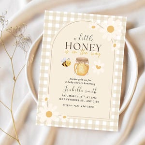 Peut inclure: Invitation de baby shower avec un fond beige à carreaux et des motifs floraux. Le texte "a little HONEY is on the way" est affiché au-dessus d'un pot de miel et d'une illustration d'abeille. L'invitation comprend les détails de la baby shower d'Isabella Smith.