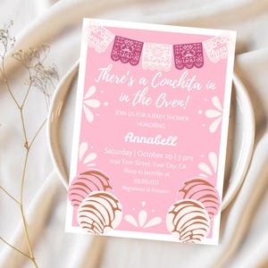 May include: Pink baby shower invitation with a white border. The invitation features a festive design with a pink background and white and pink paper cutouts. The text reads "There's a Conchita in the Oven!" and includes the name "Annabell" and details for the event.