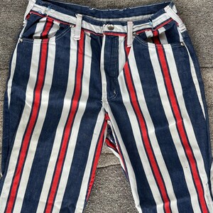 May include: Close-up of denim pants with vertical stripes in navy blue, white, and red. The pants have a classic five-pocket design with a button and zipper closure. The stripes run the length of the pants, creating a bold, eye-catching pattern.